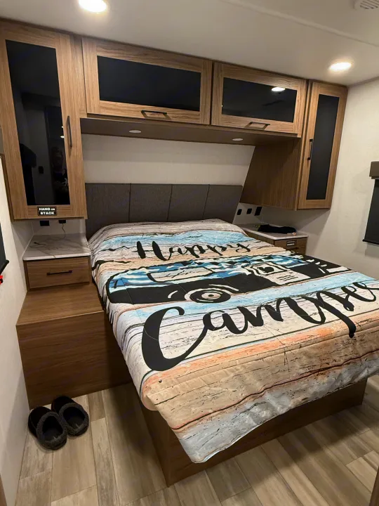 Cozy RV rental bedroom with "Happy Camper" bedding, built-in storage, and convenient nightstands for travel comfort.. Alliance RV Delta Ultra Light 2025