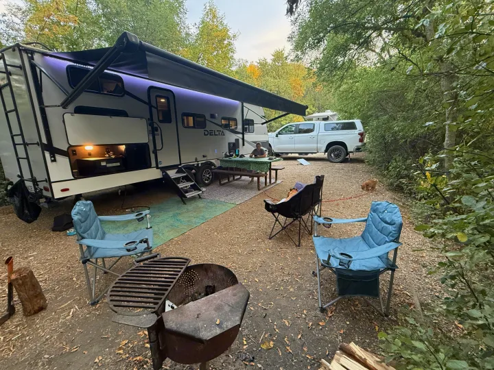 A white Delta rv rental trailer at a leafy campsite, with a picnic area, truck, camp chairs, and dogs enjoying the outdoors.. Alliance RV Delta Ultra Light 2025