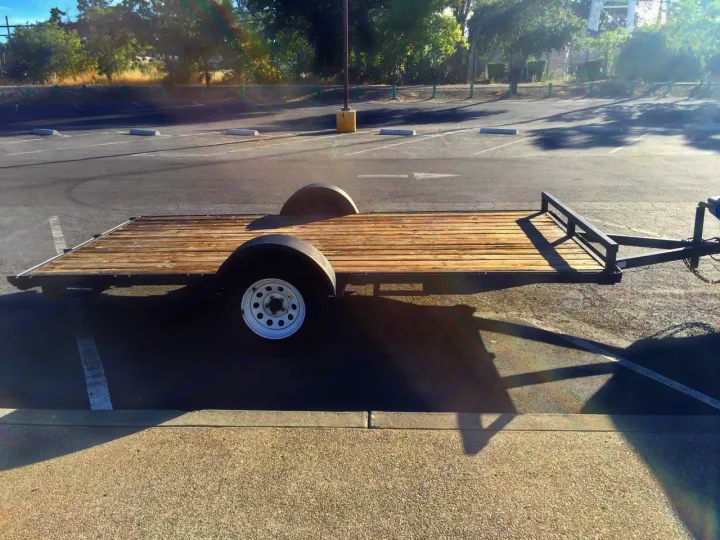 A flatbed utility trailer with wooden planks & two wheels, parked in a sunlit asphalt lot. Useful for transport with an RV rental.. Eagle 33305 2003