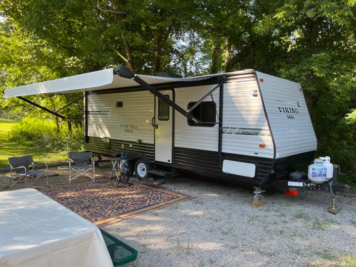 Viking Saga RV rental at a serene campsite with awning extended, chairs, rug, and picnic table, surrounded by trees.. Coachmen Viking Saga 21SBH 2019