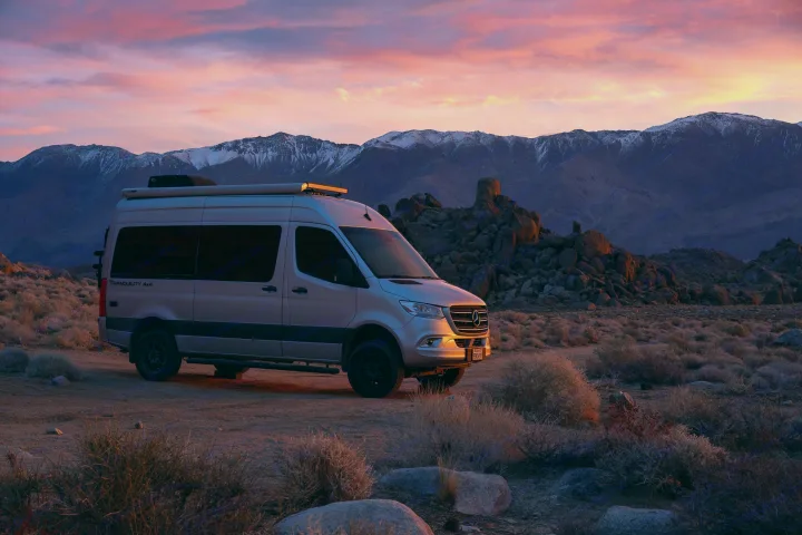 Silver Mercedes Sprinter **RV rental** at sunset. Pink sky over snow-capped mountains and a rugged desert landscape.. Thor Tranquility 2023