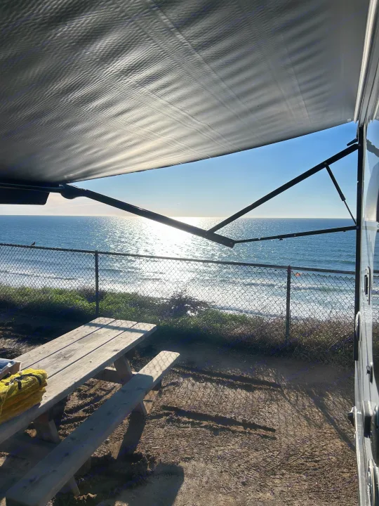 Coastal view from an RV rental with an extended awning, overlooking the sparkling ocean and a picnic table on the dirt ground.. Forest River Cherokee Wolf Pup 2023