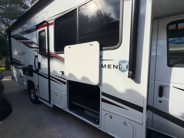 Side view of a white Coachmen Leprechaun RV rental with an open outdoor TV compartment and black/red graphics.. Coachmen Leprechaun 2025
