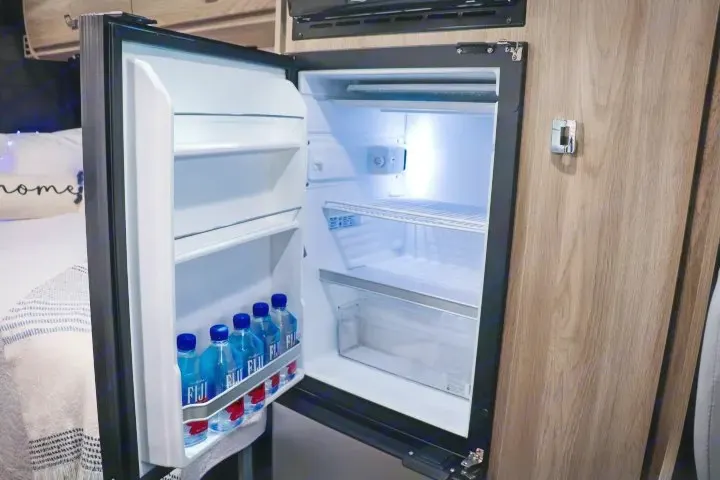 This RV rental mini-fridge, brightly lit and open, displays shelves of Fiji water bottles. White interior, wood cabinetry surrounds.. Winnebago Travato 2022