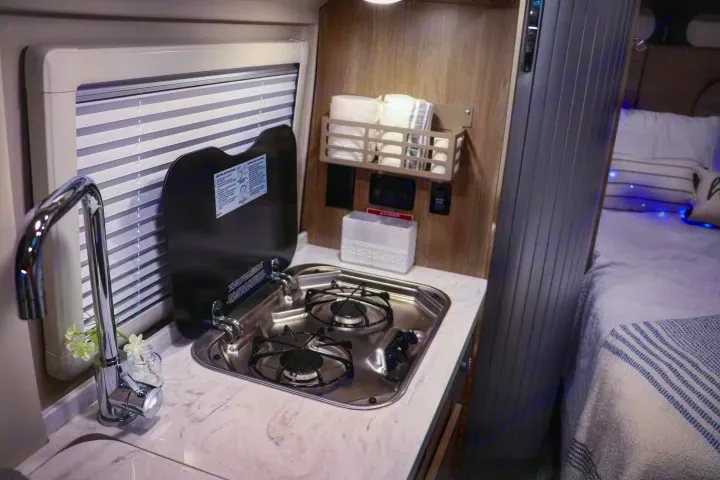 This RV rental features a compact kitchen with a two-burner stove, sink, and window. A cozy bed is visible, perfect for on-the-go living.. Winnebago Travato 2022