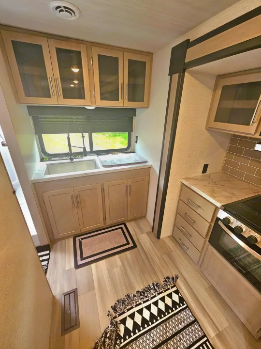 Modern RV rental kitchen featuring light wood cabinets, a sink under a window, and a stove. Perfect for your travels.. Keystone RV Springdale 2024