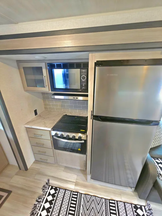 Compact RV rental kitchen features stainless steel appliances, marble-look counter, gray subway tile backsplash, and light wood flooring.. Keystone RV Springdale 2024