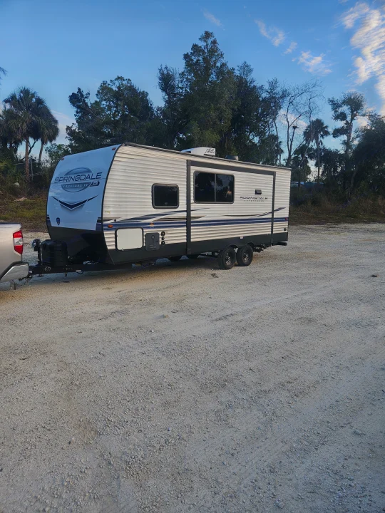 Springdale RV rental hitched to a truck on a gravel lot under a blue sky, surrounded by trees.. Keystone RV Springdale 2024