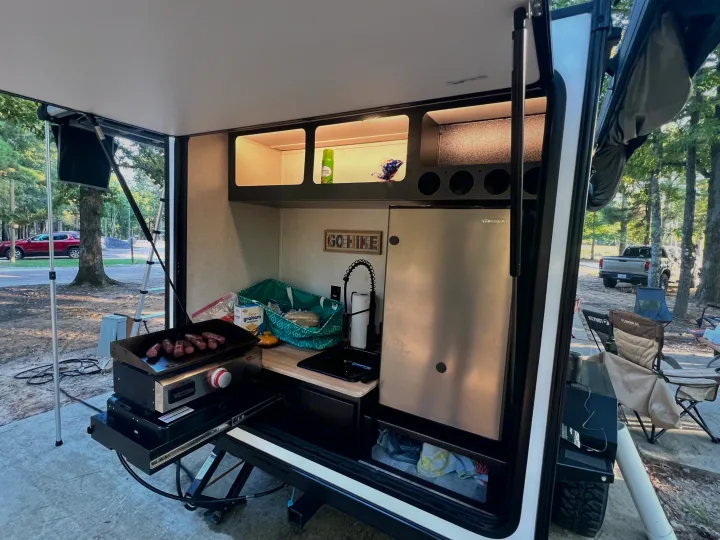 Outdoor kitchen of an RV rental, complete with a griddle cooking sausages, sink, fridge, and shelves for camping.. Forest River NoBo 10.7 2024