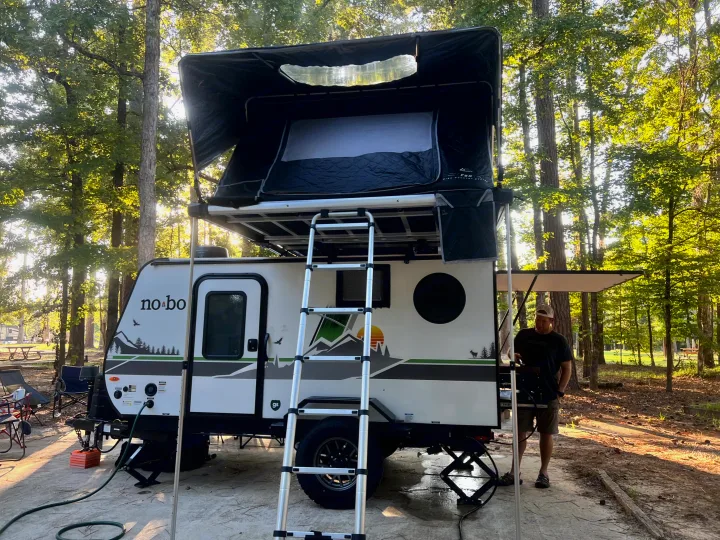 Enjoying a no-bo RV rental with a rooftop tent at a sunny campsite, complete with ladder access. A man stands nearby.. Forest River NoBo 10.7 2024