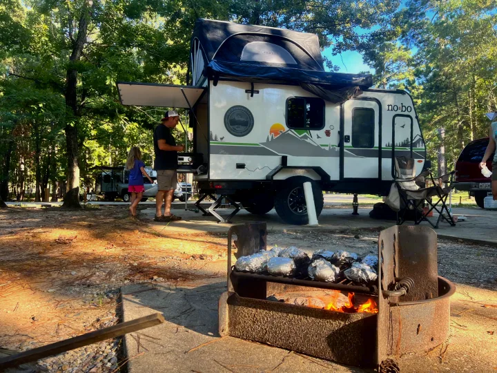 Family camping fun with an RV rental! Cooking foil-wrapped food over a campfire next to a "No Boundaries" trailer.. Forest River NoBo 10.7 2024