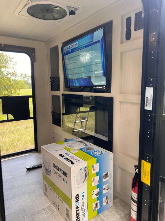 Inside an RV rental, a TV, electric fireplace, and a boxed Thetford Porta Potti 345 sit near the open door.. Forest River NoBo 10.7 2024