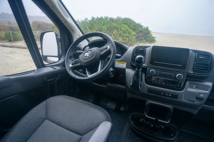 RV rental camper van interior: driver's view of dashboard, steering wheel, and a sandy, foggy beach outside.. Winnebago Solis 2025