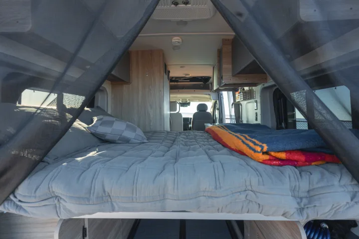 RV rental: Cozy camper van interior featuring a comfy grey bed with a colorful blanket, looking through netting to the front cab.. Winnebago Solis 2025