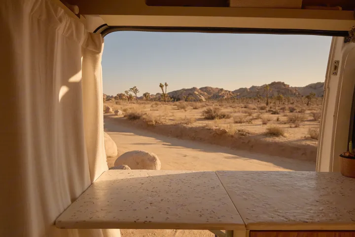 Inside an RV rental, a folding counter and curtains frame a window overlooking a desert road, Joshua trees, and distant mountains.. Dodge Ram 1500 2021