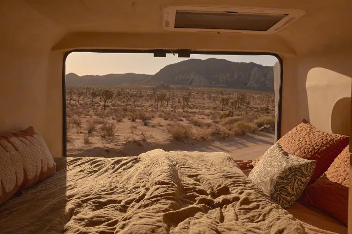 Inside an rv rental: a comfy bed with warm blankets, framing a window view of a desert, Joshua trees, & mountains.. Dodge Ram 1500 2021