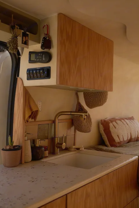 Warm rv rental interior: custom kitchen with brass faucet, speckled counter, control panel, and adjacent cozy bed.. Dodge Ram 1500 2021