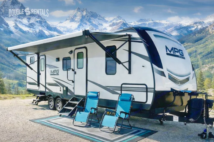 Explore the Rockies with an rv rental! Modern travel trailer with awning, chairs, and rug, set against snowy mountains.. Cruiser RV MPG 2023