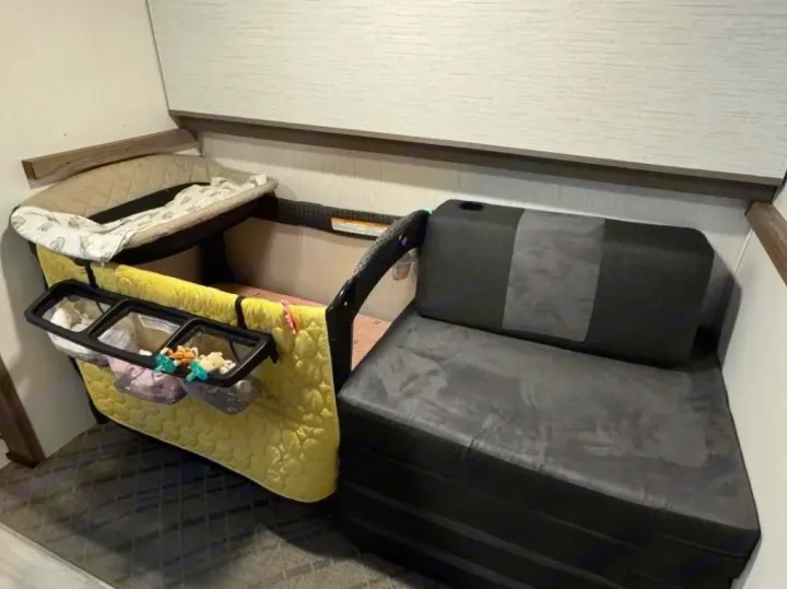 Compact RV rental space with baby playard, bassinet, and attached organizer next to a small dark sofa with a cup holder.. Cruiser RV MPG 2023