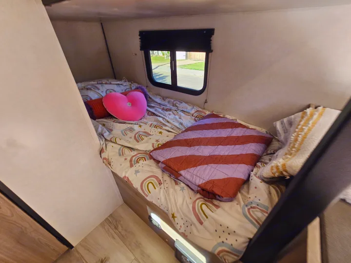 Cozy bunk bed with rainbow sheets, a heart pillow, and a window view in an RV rental. Ideal for kids' travel.. Keystone Coleman 17BX 2025