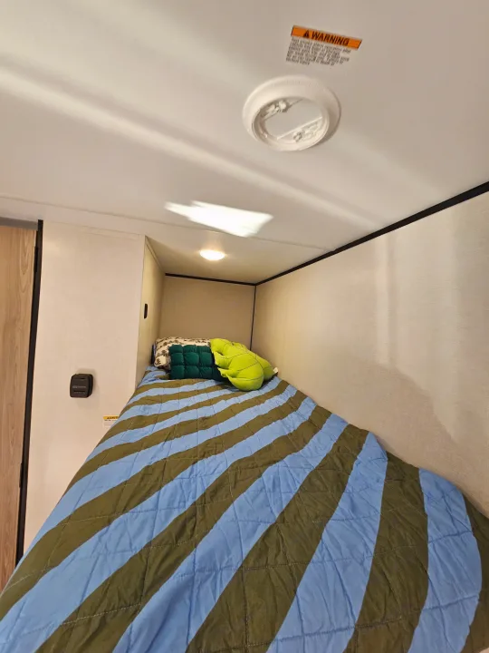 Cozy RV rental bunk bed with striped comforter & pillows. Features a thermostat, ceiling light, and warning sign.. Keystone Coleman 17BX 2025