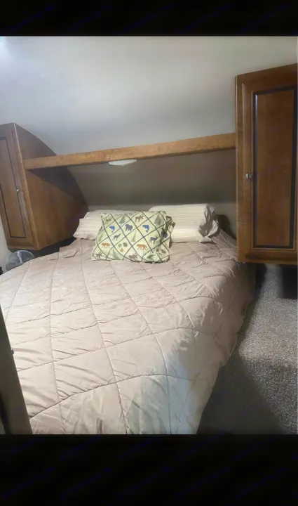A compact RV bedroom shows a quilted bed with an animal-print pillow, wood overhead cabinets, and a fan, ready for your rv rental.. Crossroads RV Z-1 2017