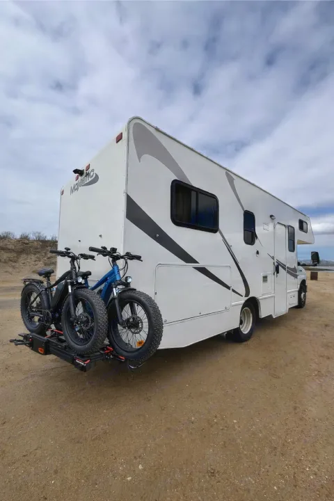 White Majestic RV rental parked outdoors, featuring a bike rack with two fat-tire electric bikes (black & blue). Cloudy sky.. Thor Majestic 2018