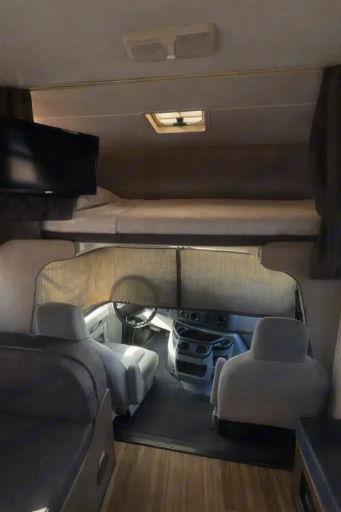 View inside an rv rental: cozy over-cab bunk with TV, front cab seating, and window shades in a camper's interior.. Thor Majestic 2018