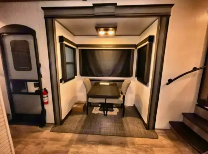 Inside an RV rental: a cozy dinette with a dark table, two upholstered chairs, windows with blinds, and nearby door & stairs.. Keystone RV Montana 2023