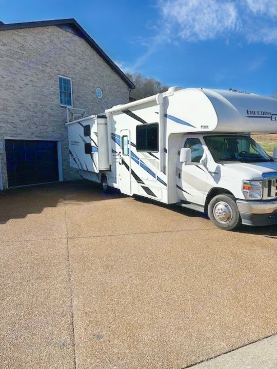 This Freedom Elite Class C RV rental, with its slide-out extended, is parked on a driveway beside a brick house on a sunny day.. Thor Freedom Elite 2021