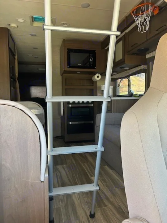 RV rental interior with a ladder leading to a bunk, a kitchen featuring a microwave, stove, sink, and a mini basketball hoop.. Thor Freedom Elite 2021