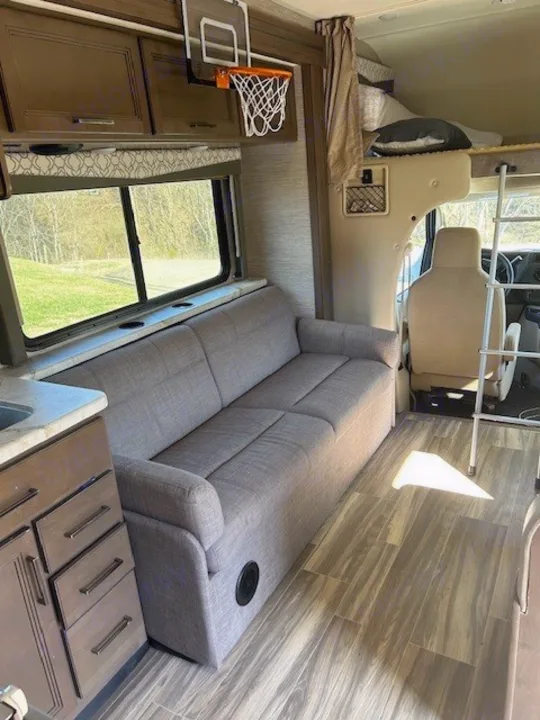 RV rental interior features a long gray sofa, kitchenette, and bunk over cab. Note the mini basketball hoop for fun on the road.. Thor Freedom Elite 2021