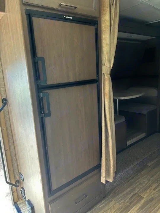 Inside this cozy RV rental, a Dometic refrigerator with wood-look panels stands next to a privacy curtain and dinette area.. Thor Freedom Elite 2021