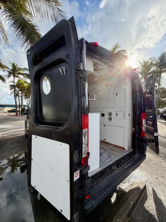 Open rear doors of a black RV rental camper van reveal its modern interior cabinetry, parked under sunny palm trees.. Gmc Ram Promaster 2018