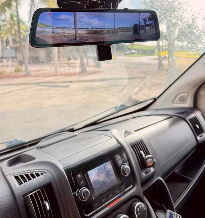 Dashboard and rain-splashed windshield of an RV rental. A digital rearview mirror shows a wide, scenic waterfront view.. Gmc Ram Promaster 2018