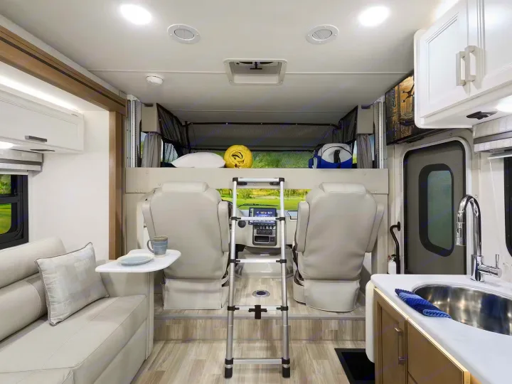 Bright RV rental interior. Features front seats, an overhead bunk bed with ladder, a living area with sofa, and a kitchen with sink.. Axis 24.1 2027