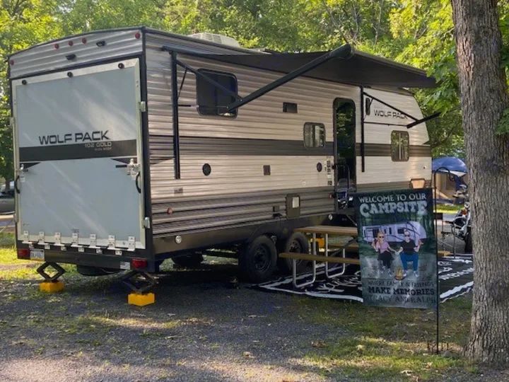 A spacious Wolf Pack RV rental at a scenic campsite, with an extended awning, picnic area, and personalized 'Welcome' flag.. Forest River Cherokee Wolf Pack 2021