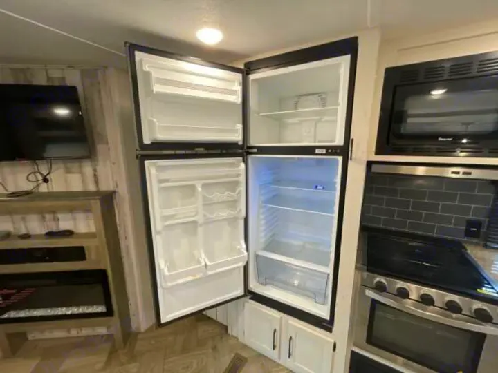 RV rental kitchen with an open two-door fridge, microwave, and stove. Adjacent is a living area with a TV and fireplace.. Palomino 28bhss 2020