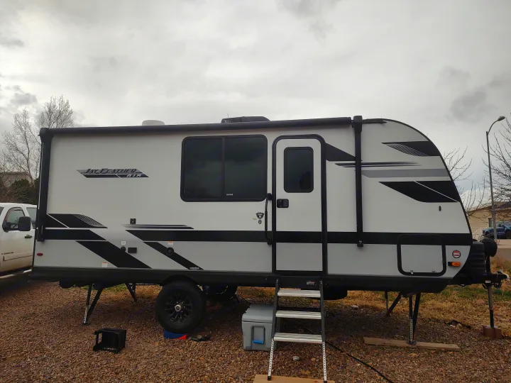 Light gray and black Jay Feather Micro RV rental, a travel trailer, parked on gravel with stabilizers down and entry ladder extended.. Jayco Jay Feather Air 18mbh 2024