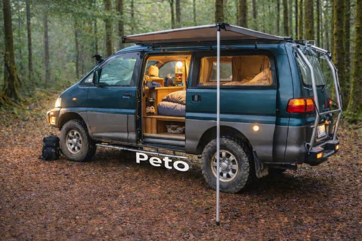Off-road campervan RV rental in a forest, side door open showing cozy lit interior with bed & storage, awning extended.. MITSUBISHI Delica 1997