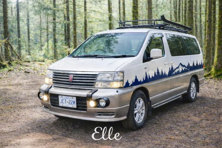 This scenic beige campervan, featuring mountain decals and a roof rack, is ideal for an RV rental wilderness adventure.. nissan elgrand 1997