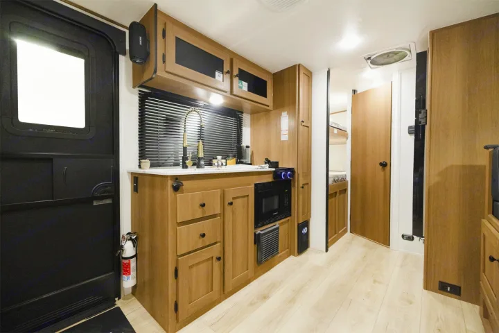 Bright rv rental interior featuring a compact kitchen with wood cabinets, sink, microwave, and an entry door. Bunk beds visible further in.. Coachmen Apex Nano 194BHS 2026