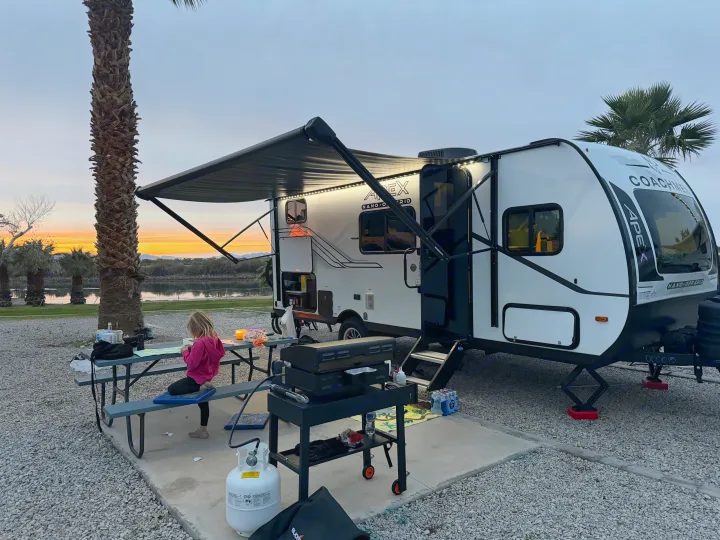 RV rental evening: Child at picnic table, grill nearby. Travel trailer parked by water with a stunning sunset backdrop.. Coachmen Apex Nano 194BHS 2026