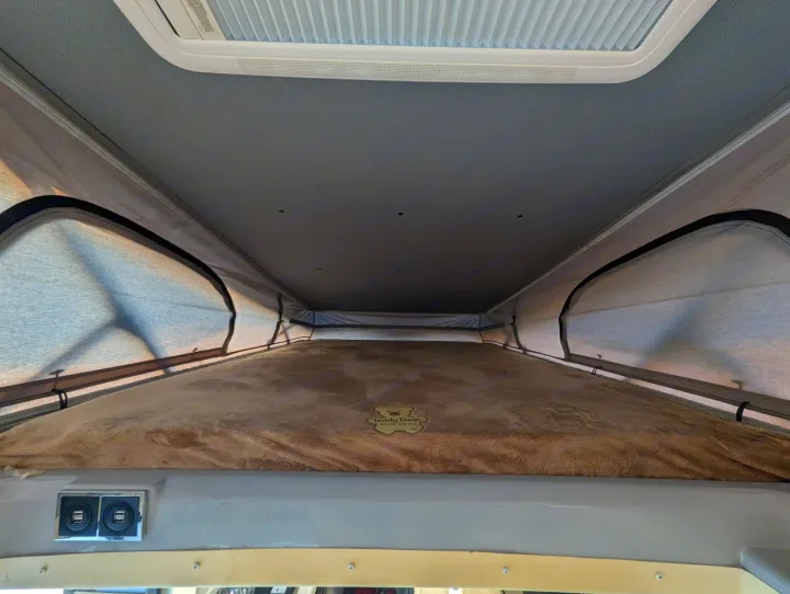 Cozy pop-top sleeping area in an RV rental, featuring a skylight, plush bed with a "Teddy Bear Bunk Series" mattress, and USB ports.. Thor Tellaro 2023
