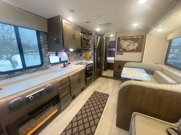 RV rental interior showcasing a modern kitchen with blue tile backsplash, cozy dining booth, and snowy window view.. Coachmen Leprechaun 2022