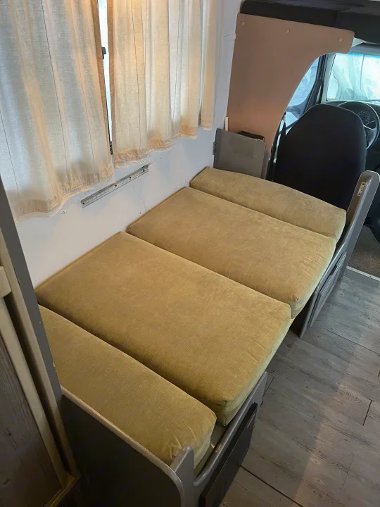 Interior of an RV rental showing a long bench seat with olive green cushions, light curtains, and wood-look flooring.. Gulf Stream Ultra 1998