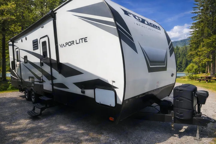 RV rental: A Fuzion Vapor Lite travel trailer is parked on gravel at a scenic campground, with a lake and trees in the background.. Keystone RV Fuzion Toy Hauler 2020