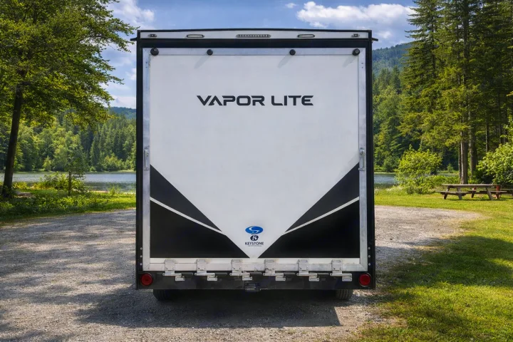 Explore nature with this Vapor Lite RV rental! Rear view shows it parked by a scenic lake with forests and a picnic area.. Keystone RV Fuzion Toy Hauler 2020
