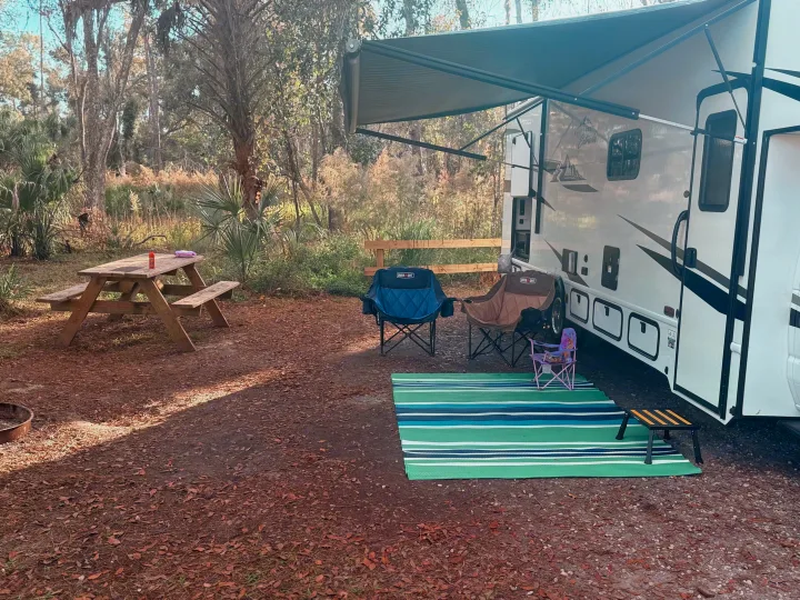 RV rental campsite setup featuring an RV with an awning, picnic table, chairs, and an outdoor mat in a natural setting.. East to West Entrada 2025