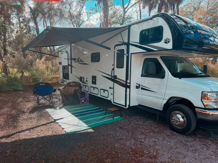 This RV rental offers a cozy campsite scene with its awning out, chairs, and outdoor mat for a relaxing outdoor experience.. East to West Entrada 2025